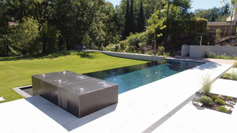 Moderner Außenpool mit dunklem Wasser, Liegefläche und umliegender grüner Gartenlandschaft